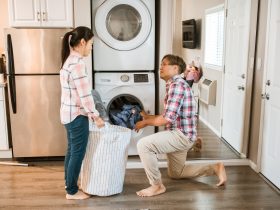 Man loading clothes in a washing machine while woman is holding a laundry basket